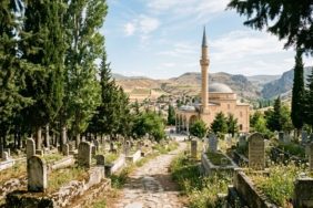 A cinematic, wide-angle shot of a serene Anatolian landscape at noon, featuring a traditional stone mosque with a single slender minaret in the background. In the foreground, a peaceful cemetery is nestled among tall, dark green cypress trees and weathered marble headstones. The warm, golden sunlight of a summer afternoon filters through the branches, casting soft shadows across the quiet grounds. The atmosphere is one of profound peace, dignity, and timeless remembrance, captured in a high-quality, editorial photography style with a soft, respectful color palette.