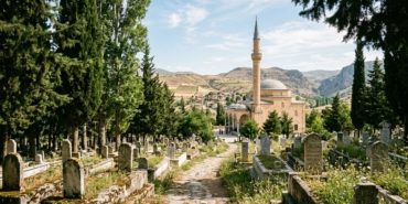 A cinematic, wide-angle shot of a serene Anatolian landscape at noon, featuring a traditional stone mosque with a single slender minaret in the background. In the foreground, a peaceful cemetery is nestled among tall, dark green cypress trees and weathered marble headstones. The warm, golden sunlight of a summer afternoon filters through the branches, casting soft shadows across the quiet grounds. The atmosphere is one of profound peace, dignity, and timeless remembrance, captured in a high-quality, editorial photography style with a soft, respectful color palette.